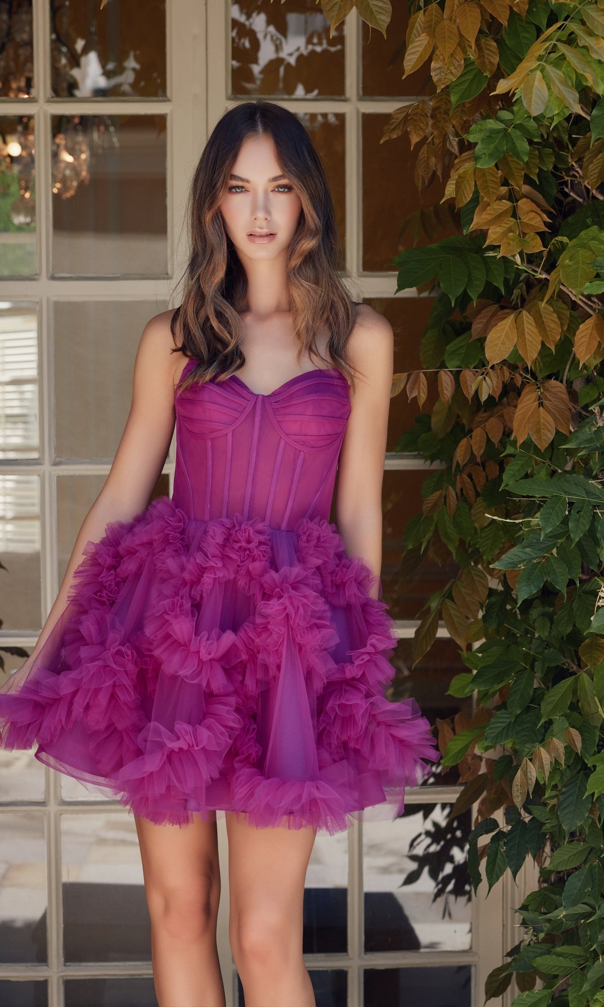 A woman in the Short Strapless Ruffled Homecoming Dress Y859, a bright magenta strapless party dress with ruffled tulle, stands before glass-paneled doors and leafy green vines.